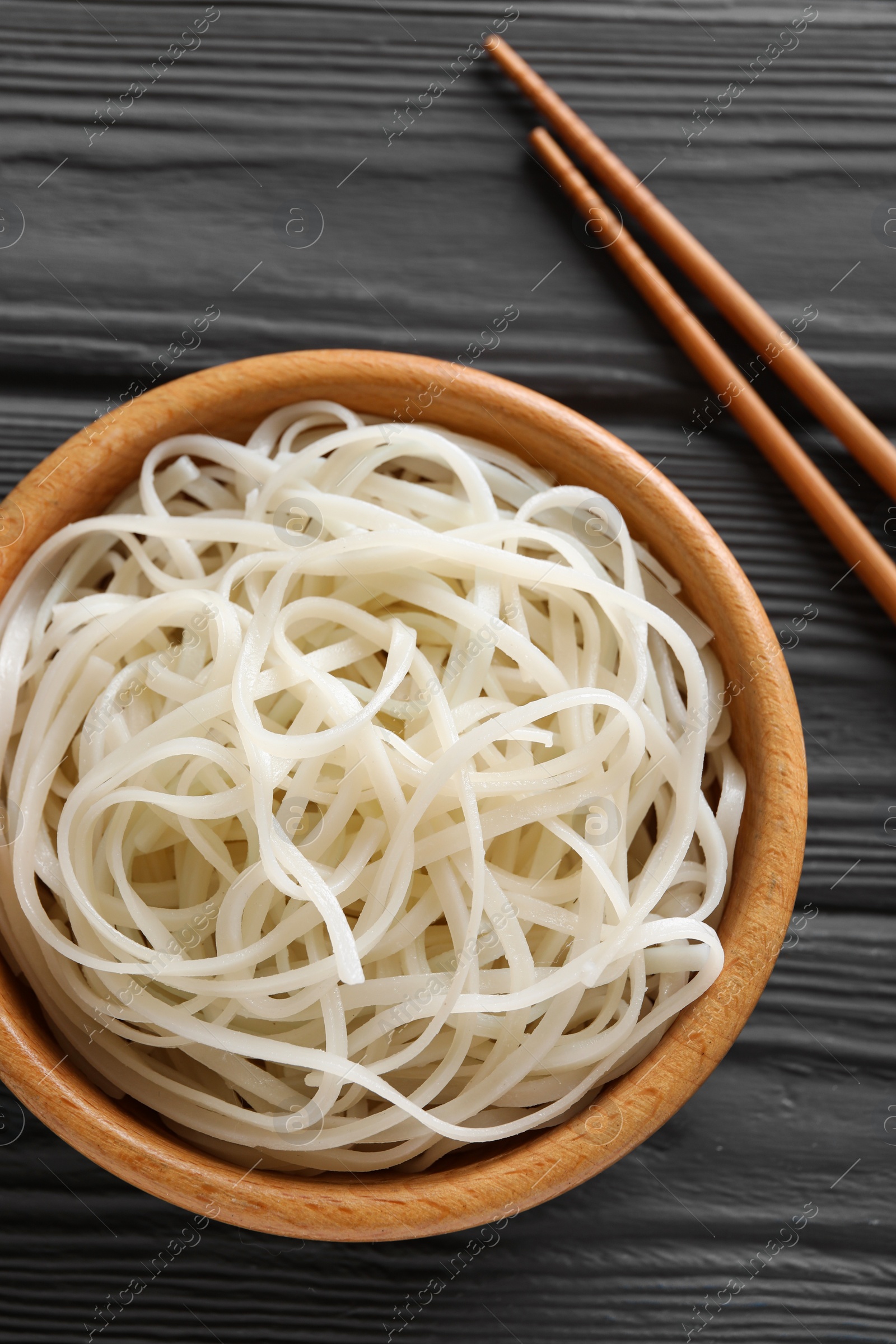 Photo of Bowl of tasty cooked rice noodles and chopsticks on black wooden table, flat lay