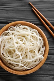 Photo of Bowl of tasty cooked rice noodles and chopsticks on black wooden table, flat lay