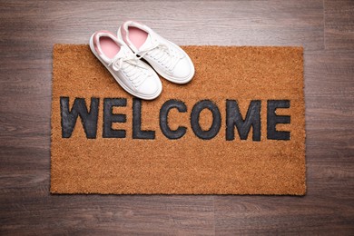 Photo of Stylish door mat with word Welcome and shoes on wooden floor, top view