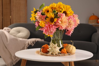 Photo of Beautiful bouquet with bright flowers and small pumpkins on white coffee table near sofa in room. Autumn atmosphere