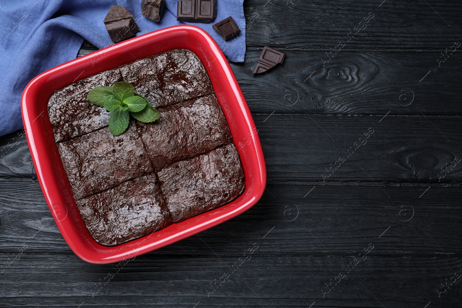 Photo of Delicious brownie with mint in baking dish and pieces of chocolate on black wooden table, flat lay. Space for text