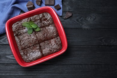 Delicious brownie with mint in baking dish and pieces of chocolate on black wooden table, flat lay. Space for text