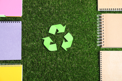 Recycling symbol, plastic and paper notebooks on green grass, flat lay
