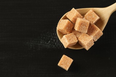 Photo of Brown sugar cubes in spoon on black wooden table, closeup. Space for text