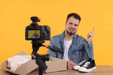 Photo of Smiling fashion blogger showing sneakers while recording video at table against orange background