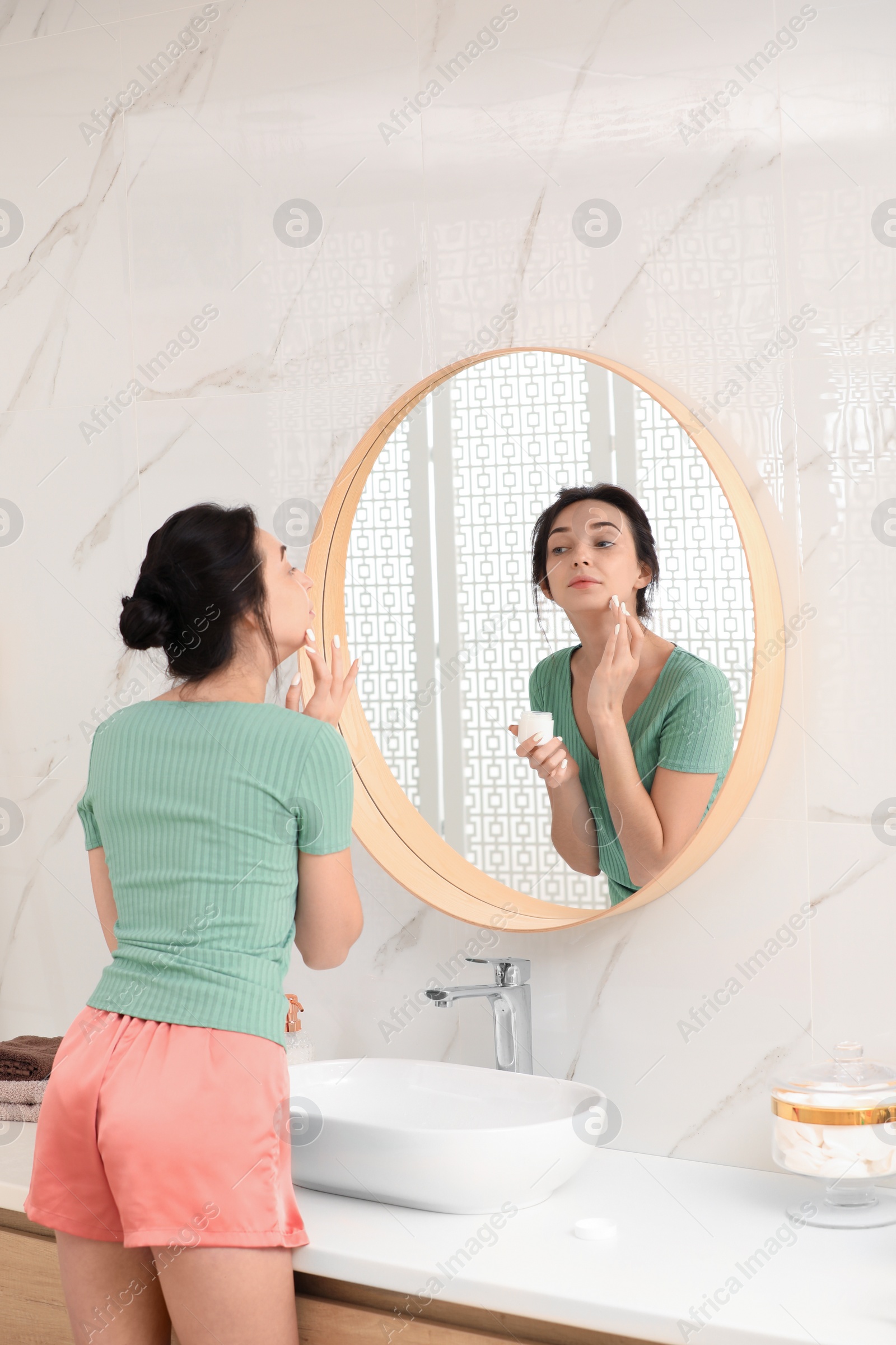 Photo of Young woman applying cream on face near mirror in bathroom
