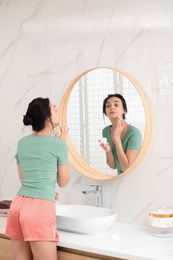 Young woman applying cream on face near mirror in bathroom