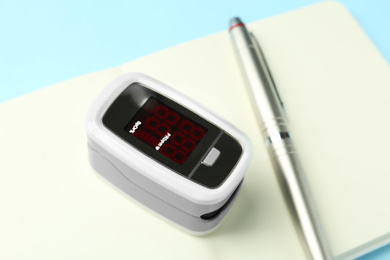 Photo of Modern fingertip pulse oximeter, pen and open notebook on light blue background, closeup