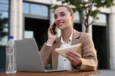 Photo of Happy businesswoman with sandwich talking on smartphone during lunch at wooden table outdoors