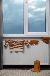 Heating radiator with knitted hat, sweater, gloves and rubber boots near window indoors
