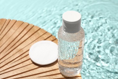 Photo of Wet bottle of micellar water and cotton pad on wooden board, closeup