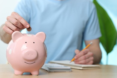 Photo of Financial savings. Man putting coin into piggy bank while writing down notes at wooden table, closeup