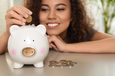 Happy African-American woman putting coin into piggy bank at table, focus on hand