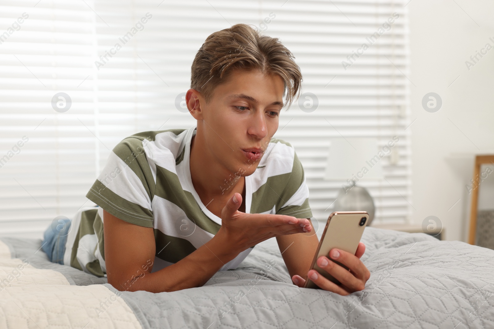 Photo of Happy young man having video chat via smartphone and blowing kiss on bed indoors. Long-distance relationship