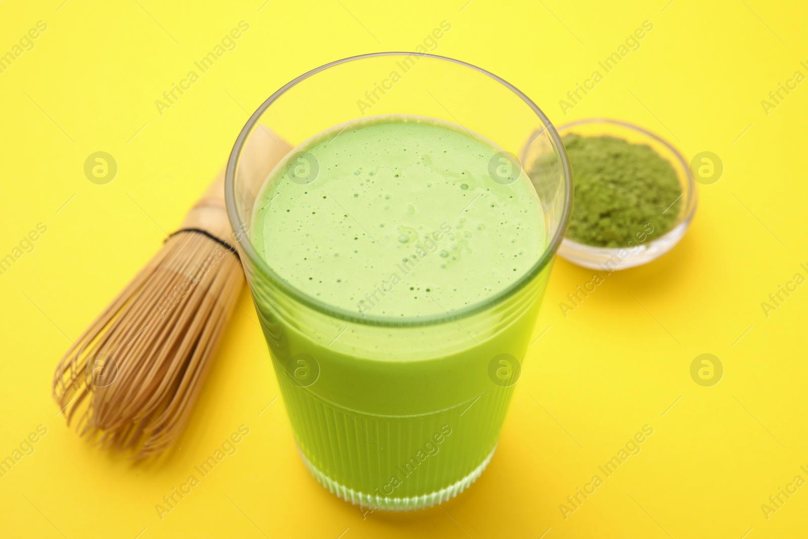 Photo of Glass of tasty matcha smoothie, powder and bamboo whisk on yellow background, closeup