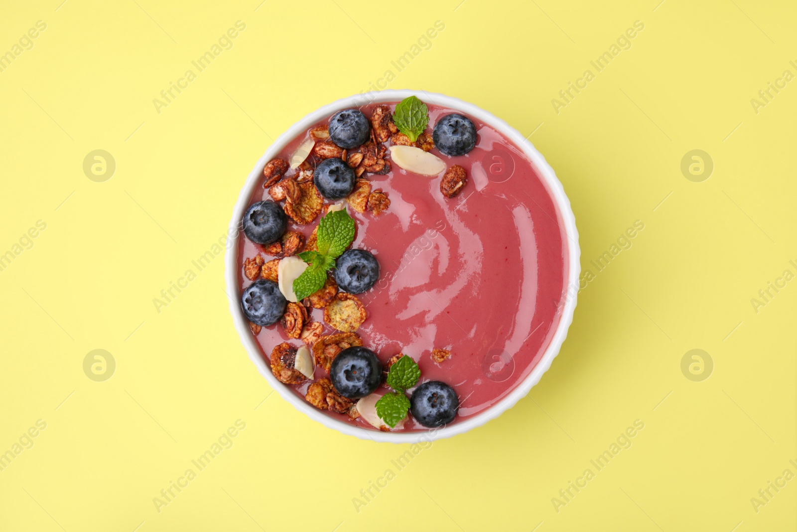 Photo of Bowl of delicious smoothie with fresh blueberries and granola on yellow background, top view