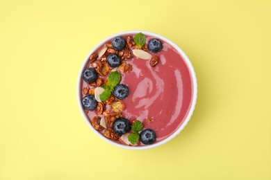 Photo of Bowl of delicious smoothie with fresh blueberries and granola on yellow background, top view