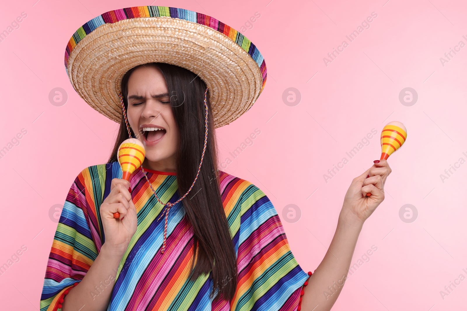 Photo of Young woman in Mexican sombrero hat and poncho singing with maracas on pink background