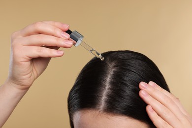 Photo of Woman applying essential oil onto hair roots on beige background, closeup