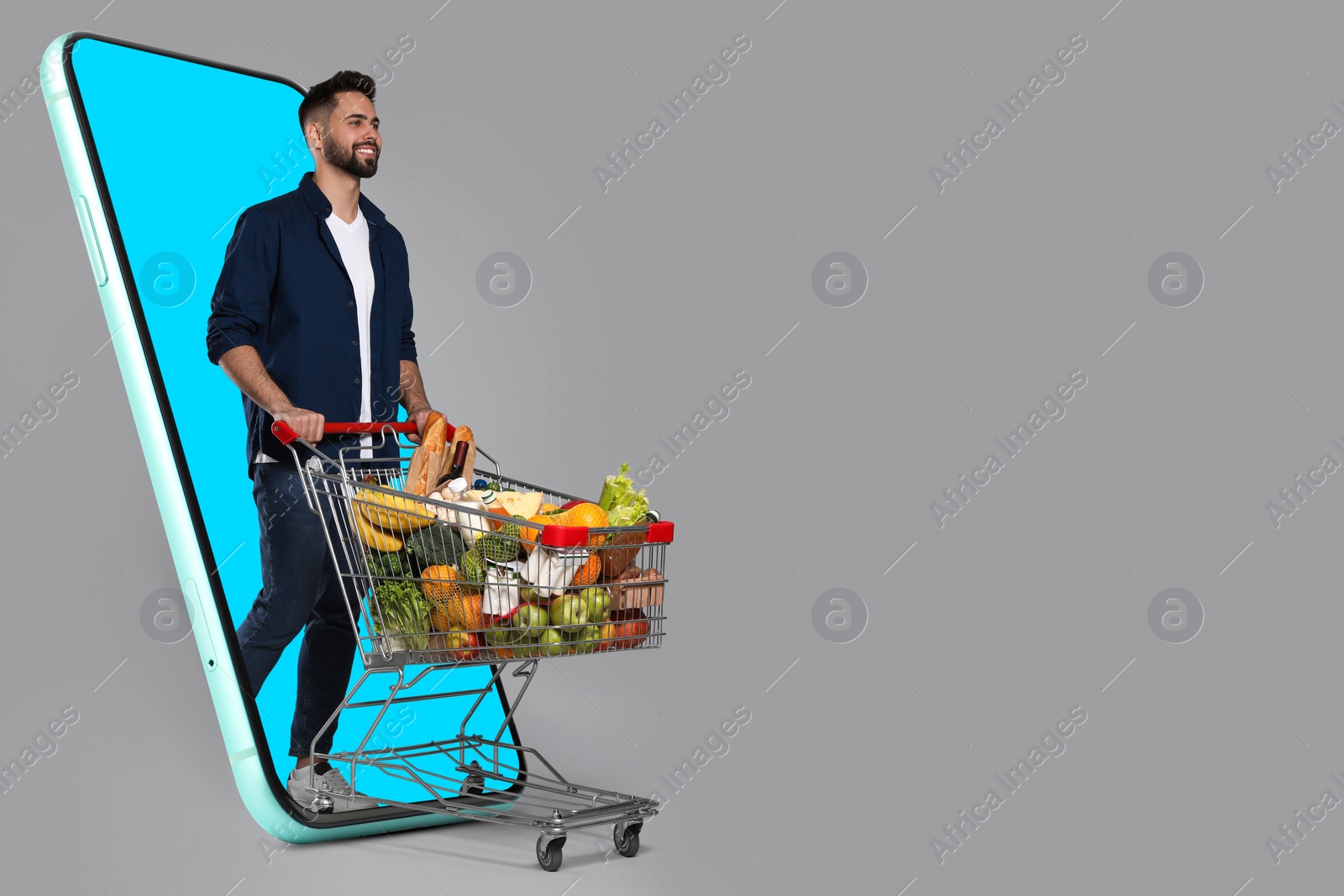 Image of Grocery shopping via internet. Happy man with shopping cart full of products walking out of huge smartphone on grey background, space for text