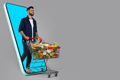 Grocery shopping via internet. Happy man with shopping cart full of products walking out of huge smartphone on grey background, space for text
