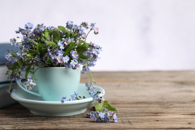 Beautiful forget-me-not flowers in cup and saucer on wooden table against light background, closeup. Space for text
