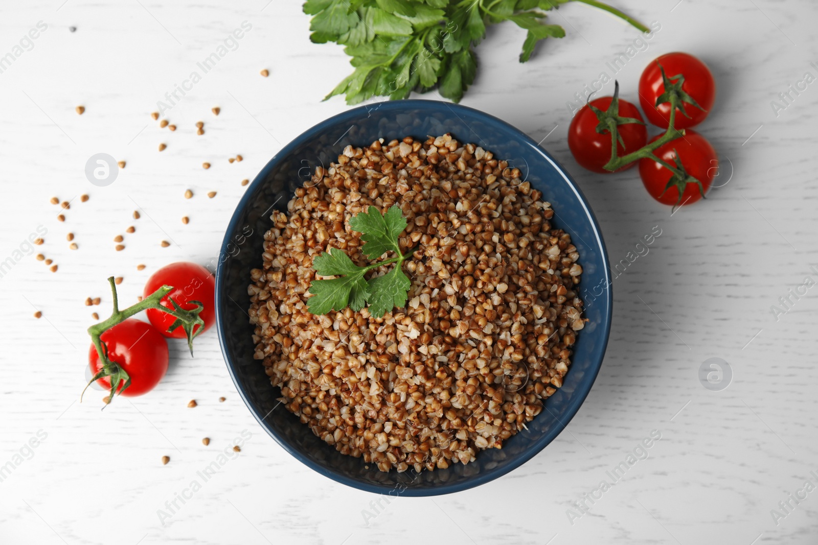 Photo of Bowl of buckwheat porridge with tomatoes and parsley on white table, flat lay