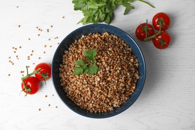 Bowl of buckwheat porridge with tomatoes and parsley on white table, flat lay