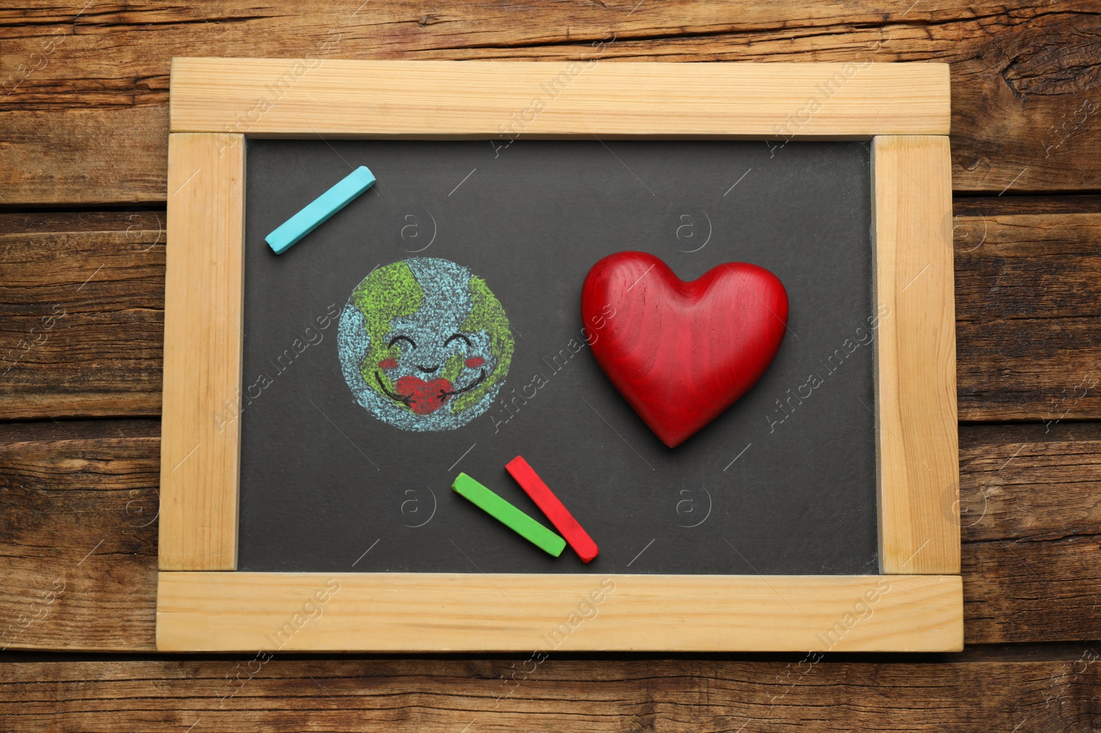 Photo of Happy Earth Day. Blackboard with drawing of planet, decorative heart and chalk pieces on wooden table, top view