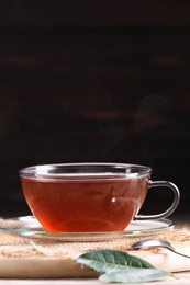 Photo of Aromatic hot tea in glass cup and leaves on wooden table against dark background. Space for text