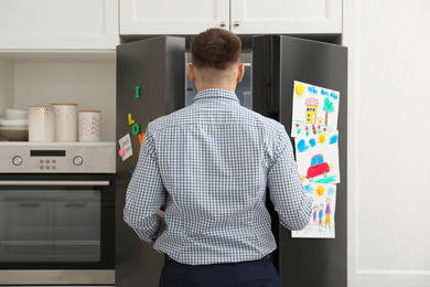 Photo of Man opening refrigerator door with child's drawings, notes and magnets in kitchen