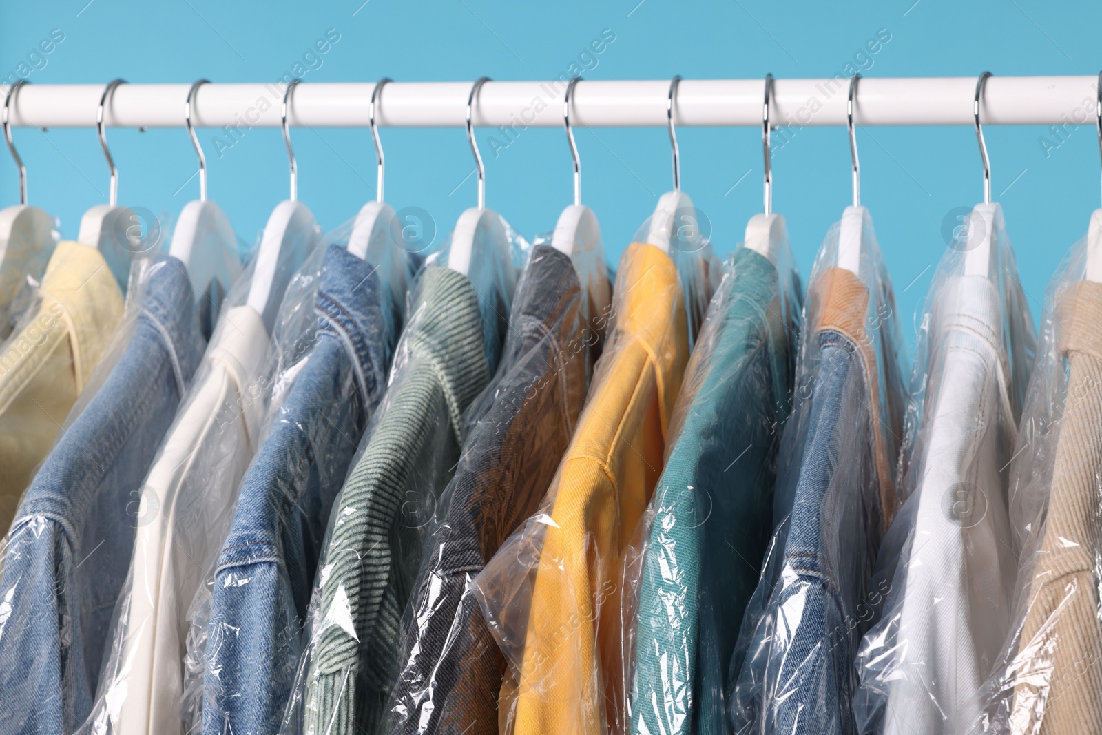 Photo of Dry-cleaning service. Many different clothes in plastic bags hanging on rack against light blue background, closeup