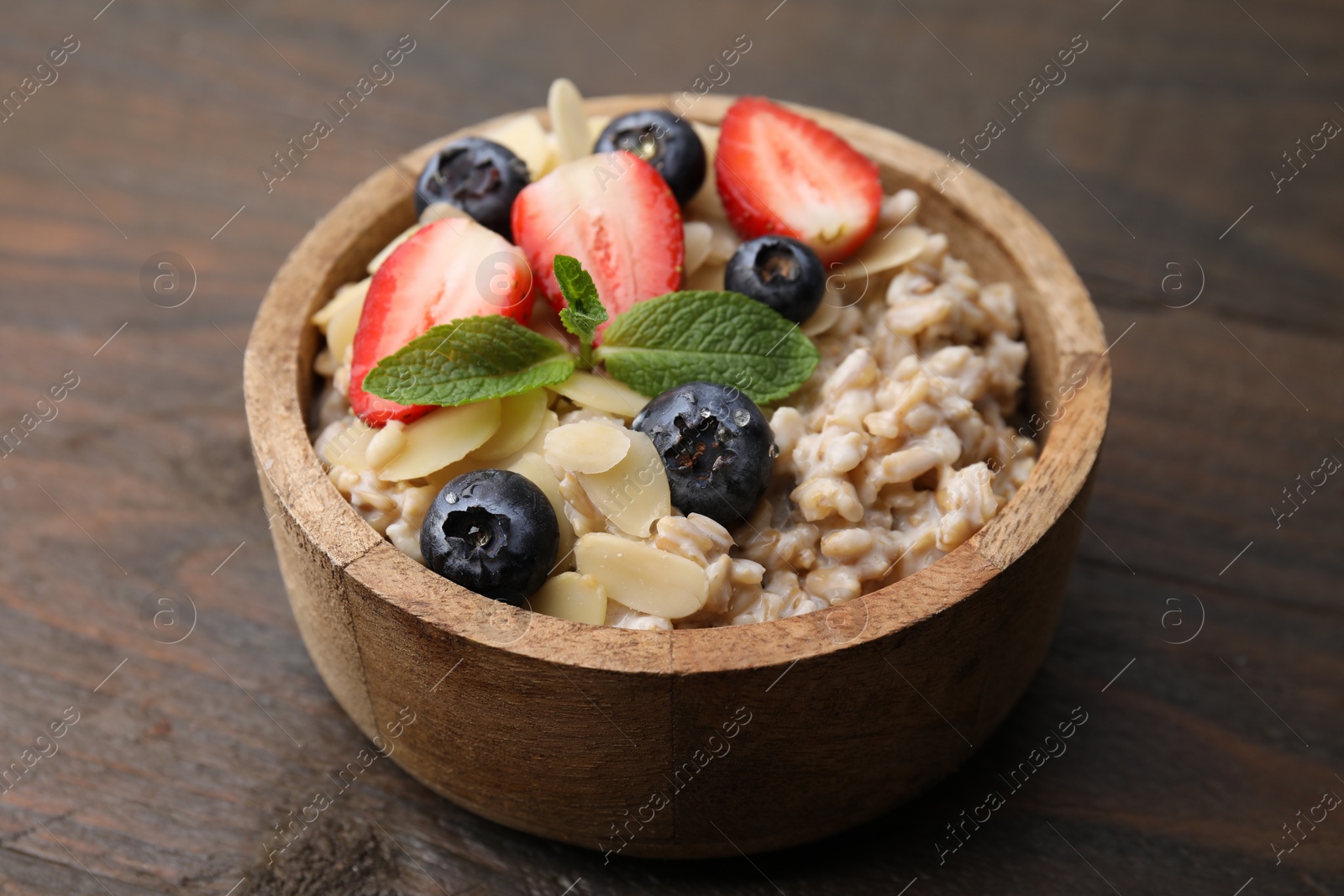 Photo of Tasty oatmeal with strawberries, blueberries and almond petals in bowl on wooden table, closeup