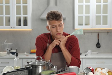 Upset man with spatula in messy kitchen. Many dirty dishware and utensils on table