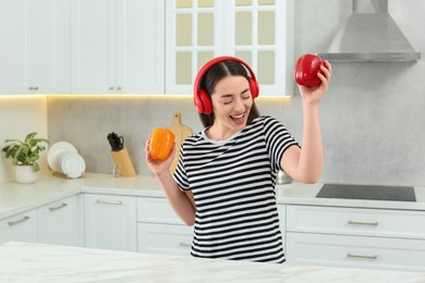 Happy woman in headphones listening music and dancing with bell peppers in kitchen