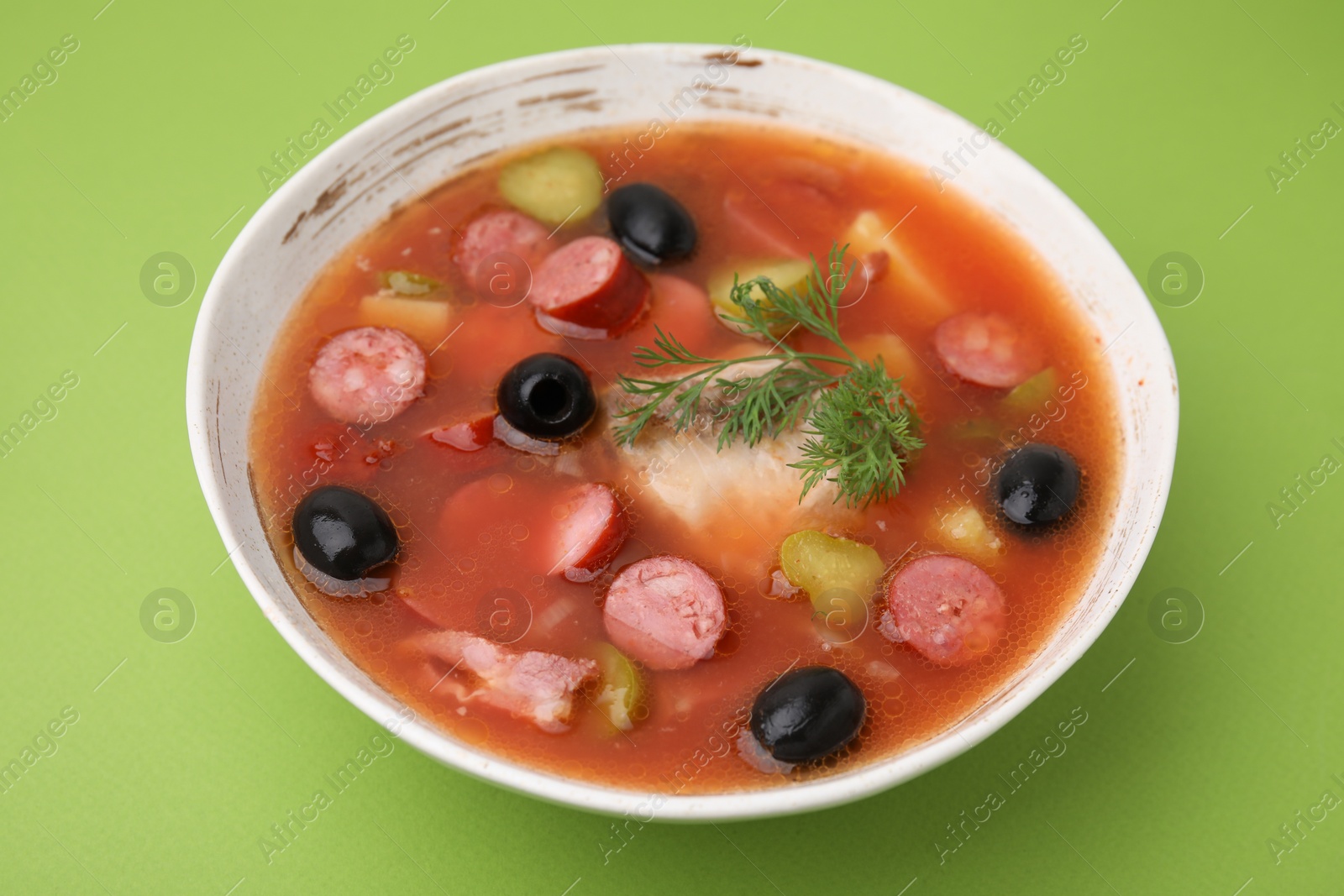 Photo of Meat solyanka soup with thin dry smoked sausages in bowl on light green background, closeup