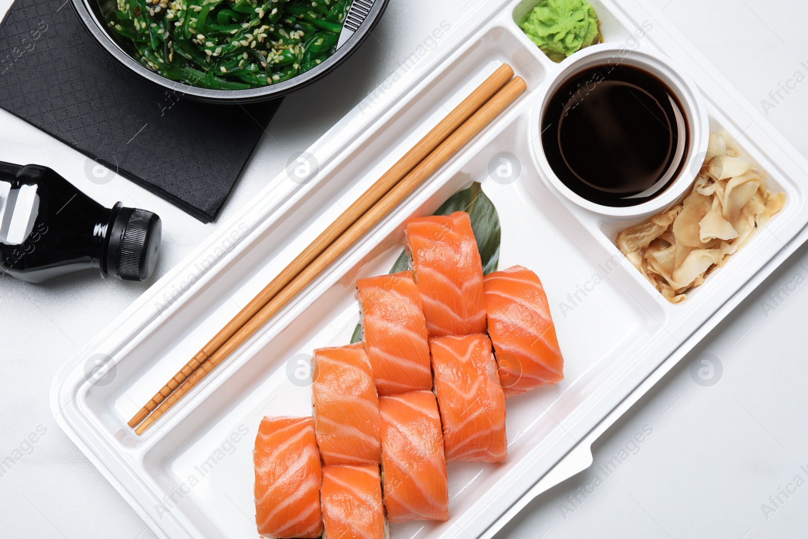 Photo of Food delivery. Delicious sushi rolls with soy sauce, ginger, wasabi and chopsticks in plastic container on white table, flat lay