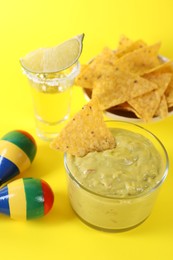 Delicious guacamole with nachos chips, maracas, tequila and lime on yellow background,