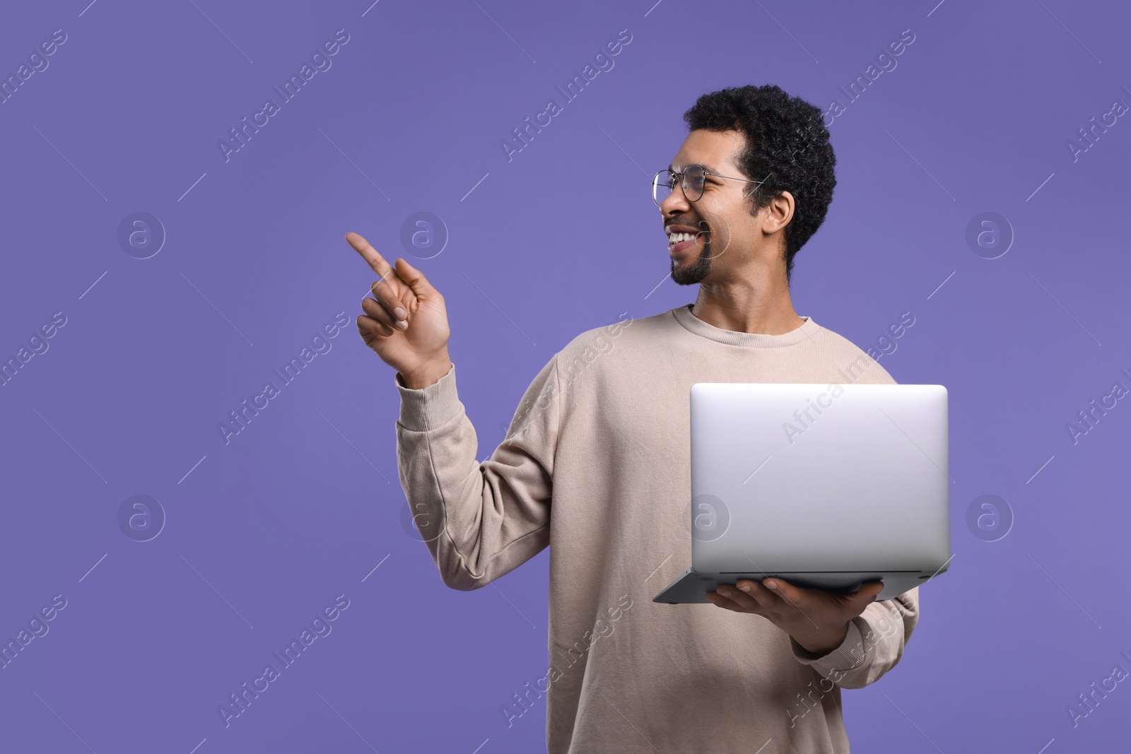 Photo of Smiling man with laptop pointing at something on purple background. Space for text