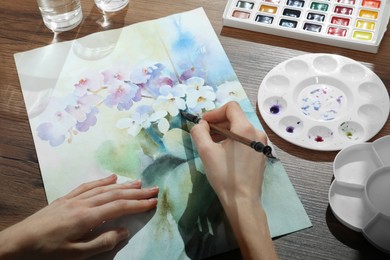 Photo of Woman painting flowers with watercolor at wooden table, closeup. Creative artwork