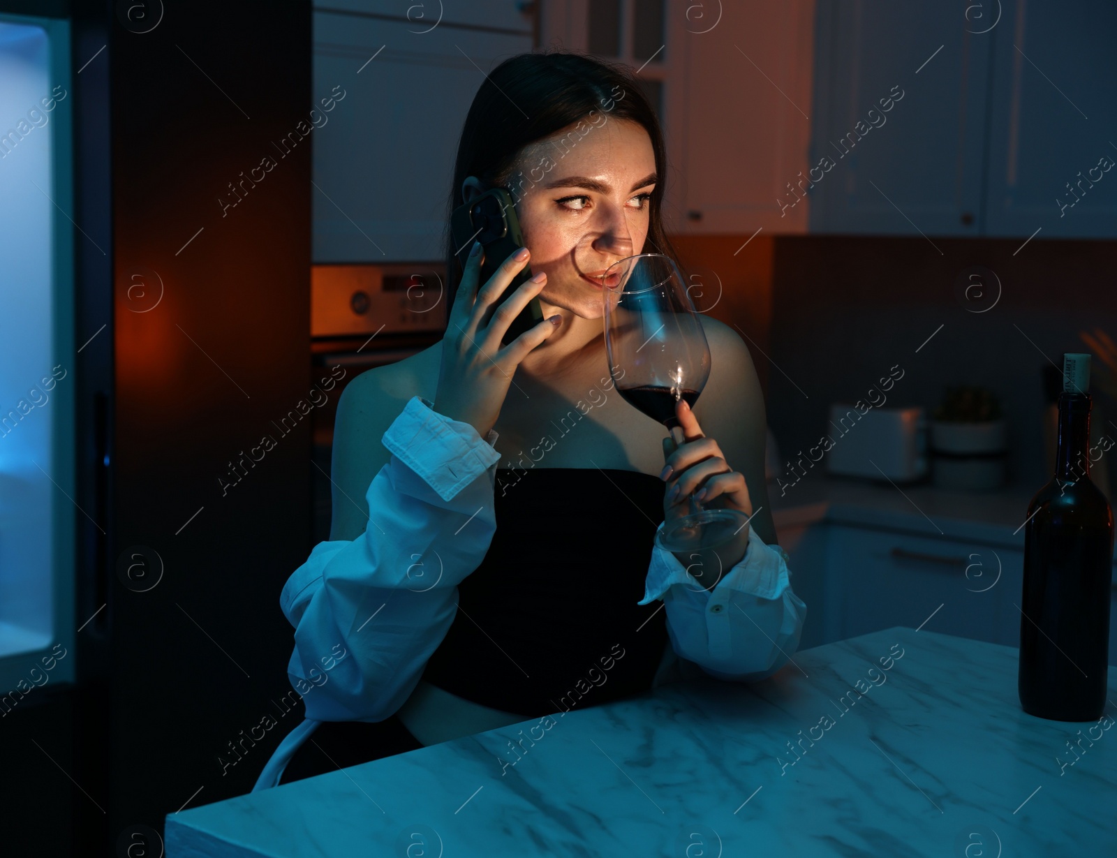 Photo of Beautiful woman talking by smartphone while drinking wine in kitchen at night