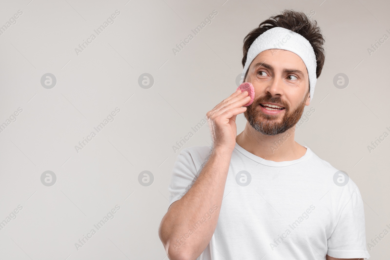 Photo of Man with headband washing his face using sponge on light grey background, space for text