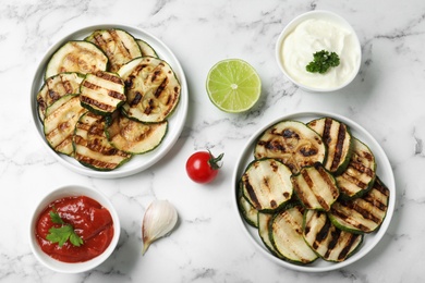 Photo of Grilled zucchini slices served with sauces on white marble table, flat lay