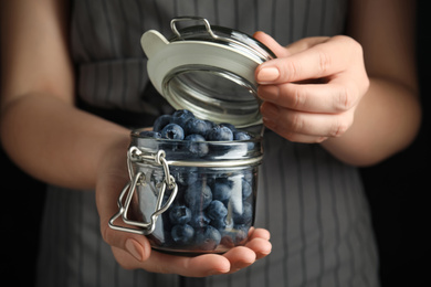 Photo of Woman holding jar with juicy fresh blueberries on black background, closeup