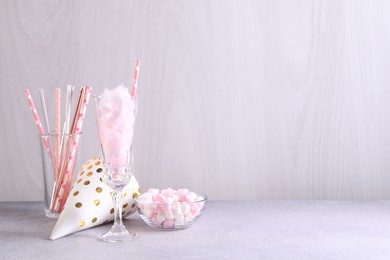 Tasty cotton candy cocktail in glass, festive decor and marshmallows on gray table against light wooden wall, space for text