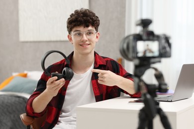 Photo of Smiling teenage blogger pointing at his headphones while streaming at home