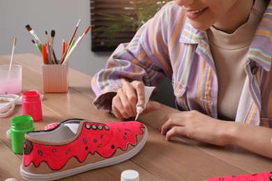 Photo of Woman painting on sneaker at wooden table indoors, closeup. Customized shoes