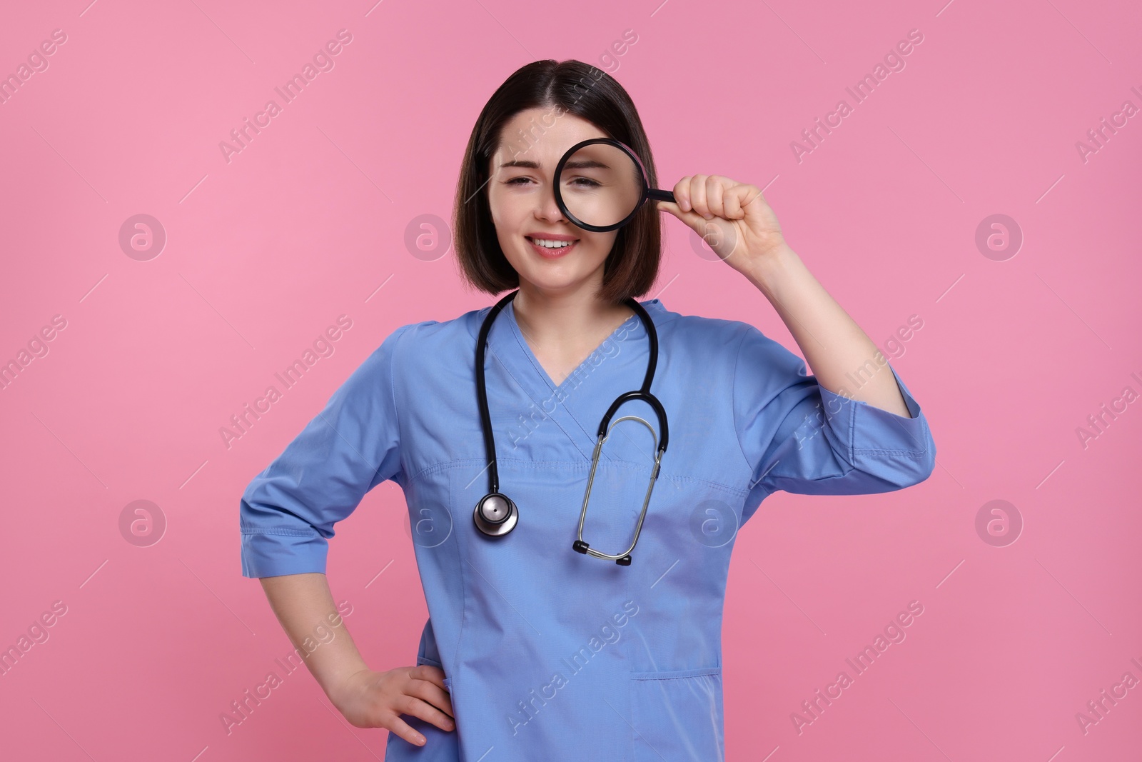 Photo of Happy young doctor with stethoscope looking through magnifier on pink background