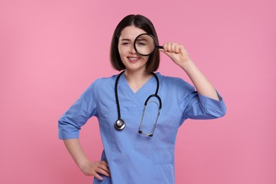 Happy young doctor with stethoscope looking through magnifier on pink background