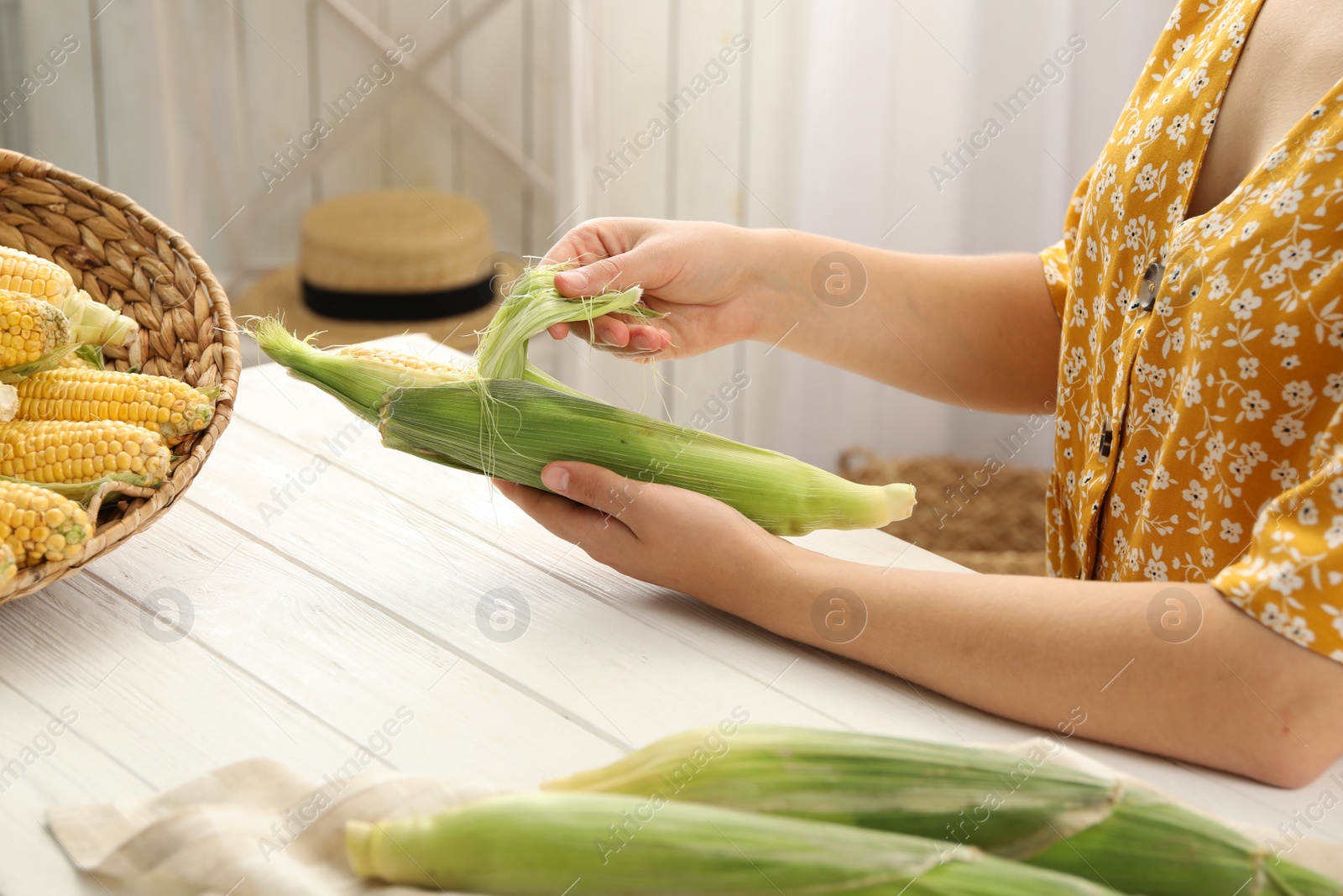 Photo of Woman husking corn cob at white wooden table, closeup